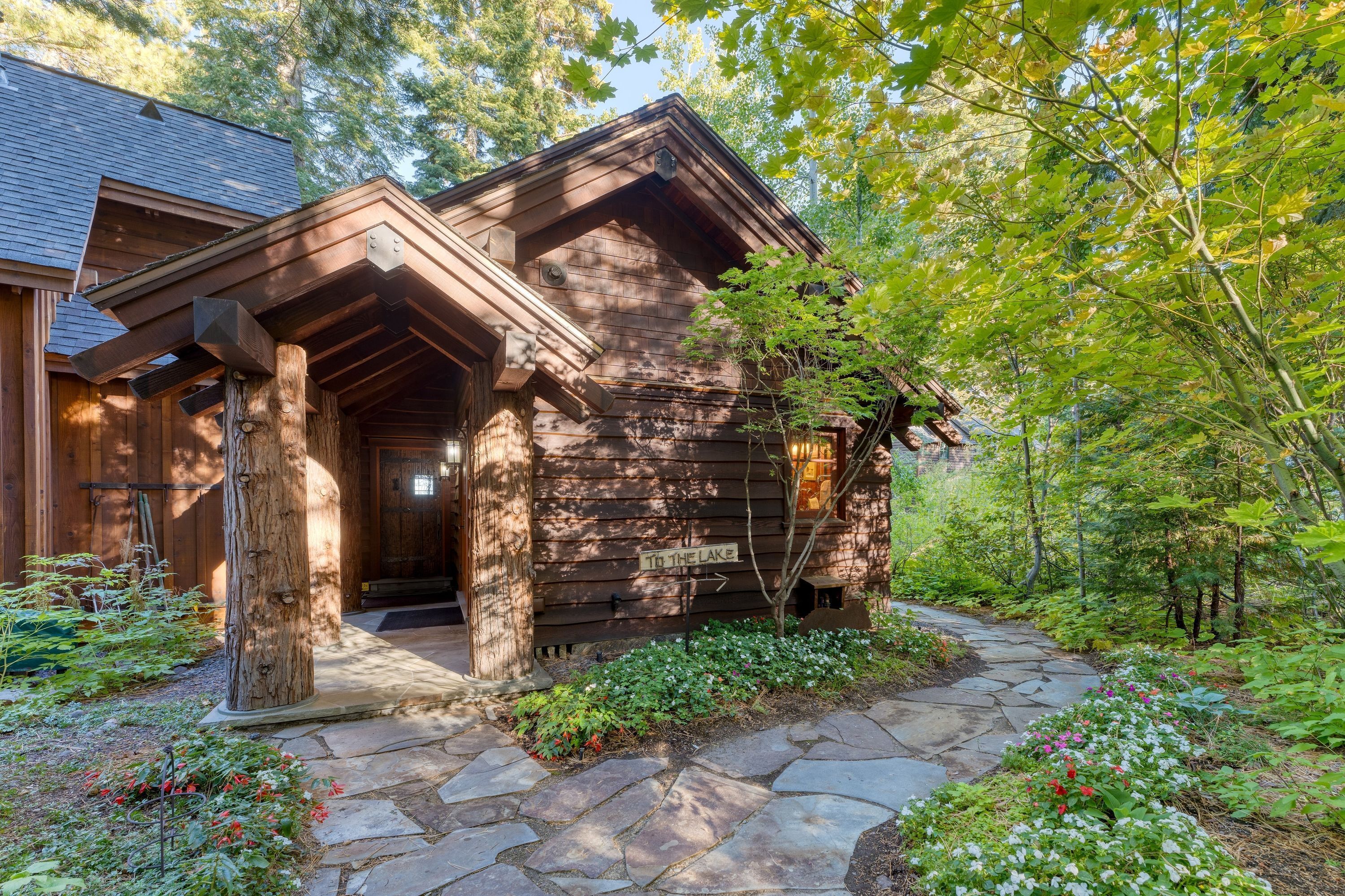 A rustic cabin with a stone pathway leading to the entrance, surrounded by lush greenery and a sign indicating a lake nearby.
