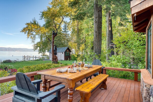 Une terrasse en bois avec une table dressée pour le repas, donnant sur un lac et entourée d’arbres.