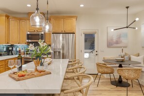 A modern kitchen with wooden cabinets, stainless steel appliances, a marble island, wicker chairs, and a dining table. - Sunset Serenity by Avantstay Rooftop w Hot Tub Fire Pit 3 Blocks to Beach (Grover Beach)