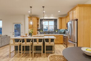 A modern kitchen with wooden cabinets, a central island with bar stools, stainless steel appliances, and a view through a window. - Sunset Serenity by Avantstay Rooftop w Hot Tub Fire Pit 3 Blocks to Beach (Grover Beach)
