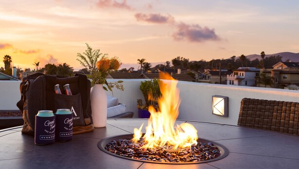 Un foyer entouré de galets, un fauteuil en osier, un vase blanc avec des plantes et deux canettes de bière sur une terrasse en toit, d’où l’on peut admirer le coucher de soleil.