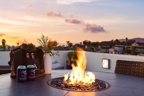 A fire feature surrounded by pebbles, a wicker chair, a white vase with plants, and two cans of beer on a rooftop terrace with a sunset view. - Sunset Serenity by Avantstay Rooftop w Hot Tub Fire Pit 3 Blocks to Beach (Grover Beach)