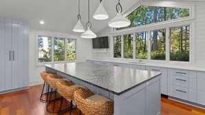 Una cocina moderna con una isla grande, taburetes de bar y ventanas amplias con vista a árboles.
