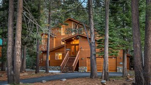 Une cabane en bois avec un escalier menant à l’entrée, entourée de grands arbres et bordée d’un chemin de pierres.