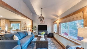 A modern living room with a large sectional sofa, a coffee table, and a television. There is a wooden ceiling, a large window with a view of trees, and a chandelier. - Washoe Way by Avantstay Luxury Tahoe Retreat A Location w Beach Access (Tahoe City)
