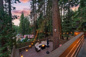 Una terraza con asientos al aire libre y luces colgantes, rodeada de árboles altos y una zona boscosa.