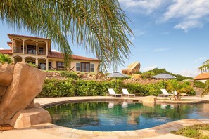 A pool area with lounge chairs, a large rock, and a house with a red roof. - Colina by Avantstay Secluded Mountain Top Oasis w Pool Hot Tub Putting Green (Murrieta)