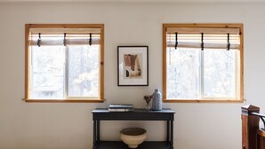 A room with two windows, a wooden console table with a bowl, a framed picture, and a vase with flowers. - Jackhorn By Avantstay Spectacular Cozy Cabin w Hot Tub (Big Bear Lake)