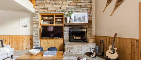 A cozy living room with a stone fireplace, wooden furniture, a guitar, and a television.