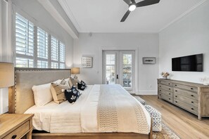 A bedroom with a bed, bedside table, lamp, dresser, TV, and large windows with blinds.