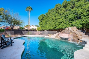 A swimming pool with a waterfall feature, surrounded by a fence and trees.