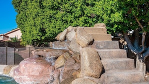 Une cascade rocheuse et des marches menant à une zone avec une piscine, le tout entouré d’arbres et avec un bâtiment en arrière-plan.