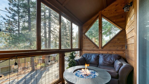 A cozy cabin interior with a fireplace, a leather sofa, and large windows offering a view of a forest. - Denton by Avantstay Hot Tub Enclosed Deck w Fire Pit Near Skiing Lake (Truckee)
