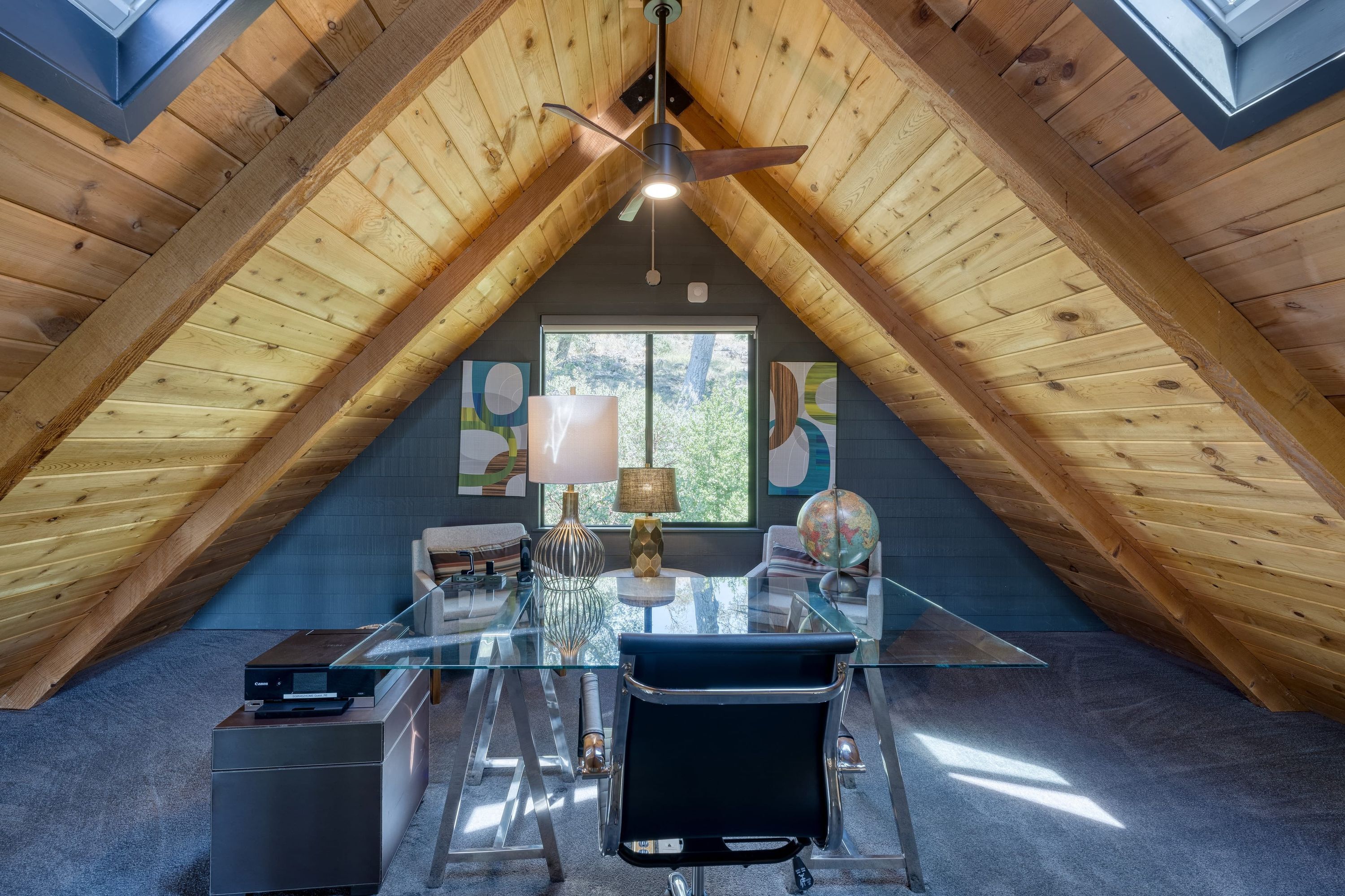 A modern attic bedroom with a glass desk, a ceiling fan, and a large window.
