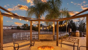 A covered outdoor seating area with a fire pit, benches, string lights, and a view of a fenced field.