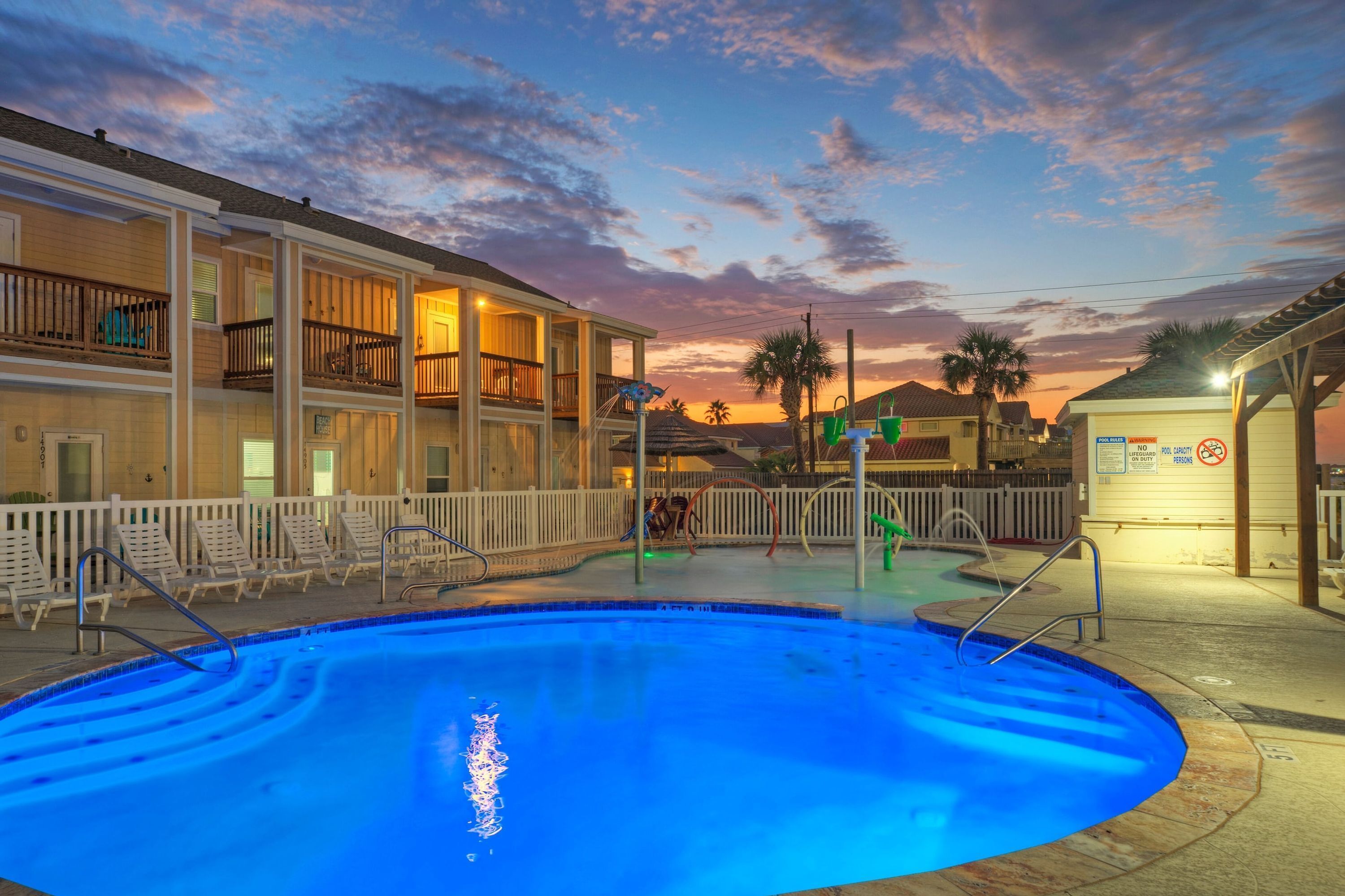 A swimming pool with a white fence, lounge chairs, and a building with balconies.