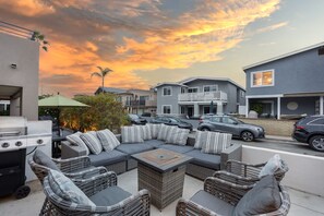 A patio area with a gray sectional sofa, wicker chairs, and a grill. - Sandy by Avantstay Oceanview Steps to Beach Patio w Fire Pit Dining Area (Newport Beach)