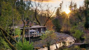 Une cabane dotée d’une terrasse donnant sur une rivière, entourée d’arbres et offrant une vue sur le coucher du soleil.