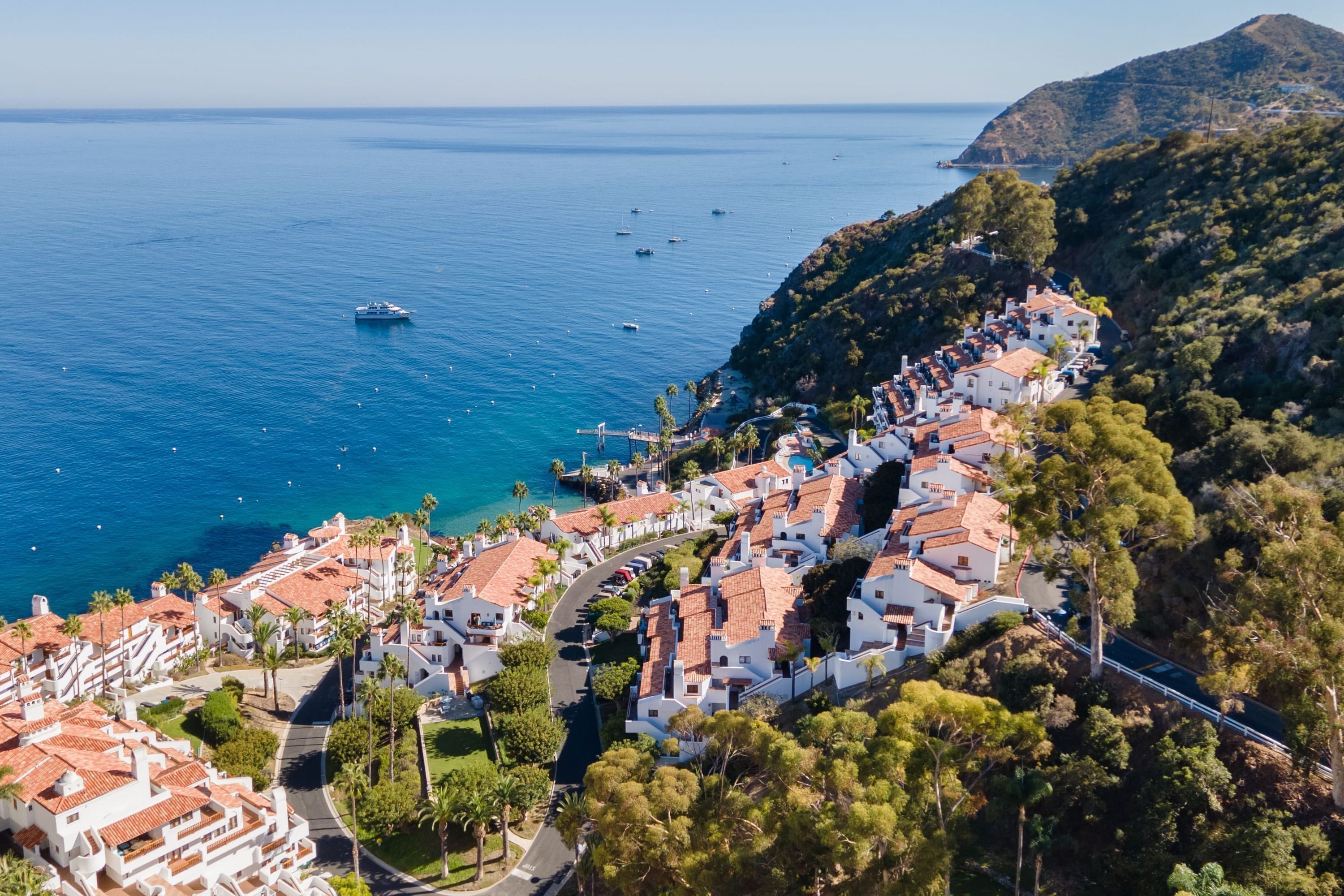 A coastal town with terracotta-roofed buildings, a marina, and a mountainous backdrop.