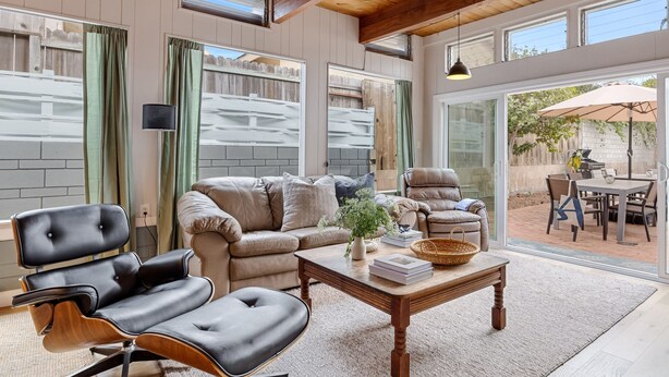 A spacious living room with a leather sofa, a matching armchair, and a wooden coffee table. Large windows provide ample natural light and a view of the backyard.