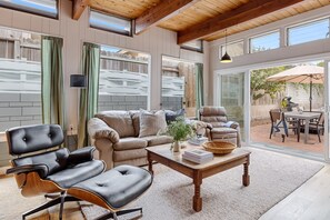 A spacious living room with a leather sofa, a matching armchair, and a wooden coffee table. Large windows provide ample natural light and a view of the backyard. - California Dreamin by Avantstay Private Pool Fire Pit Near the Beach (Carlsbad)