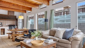 A cozy living room with a wooden ceiling, a beige sofa, a coffee table, and a dining area with a kitchen in the background.