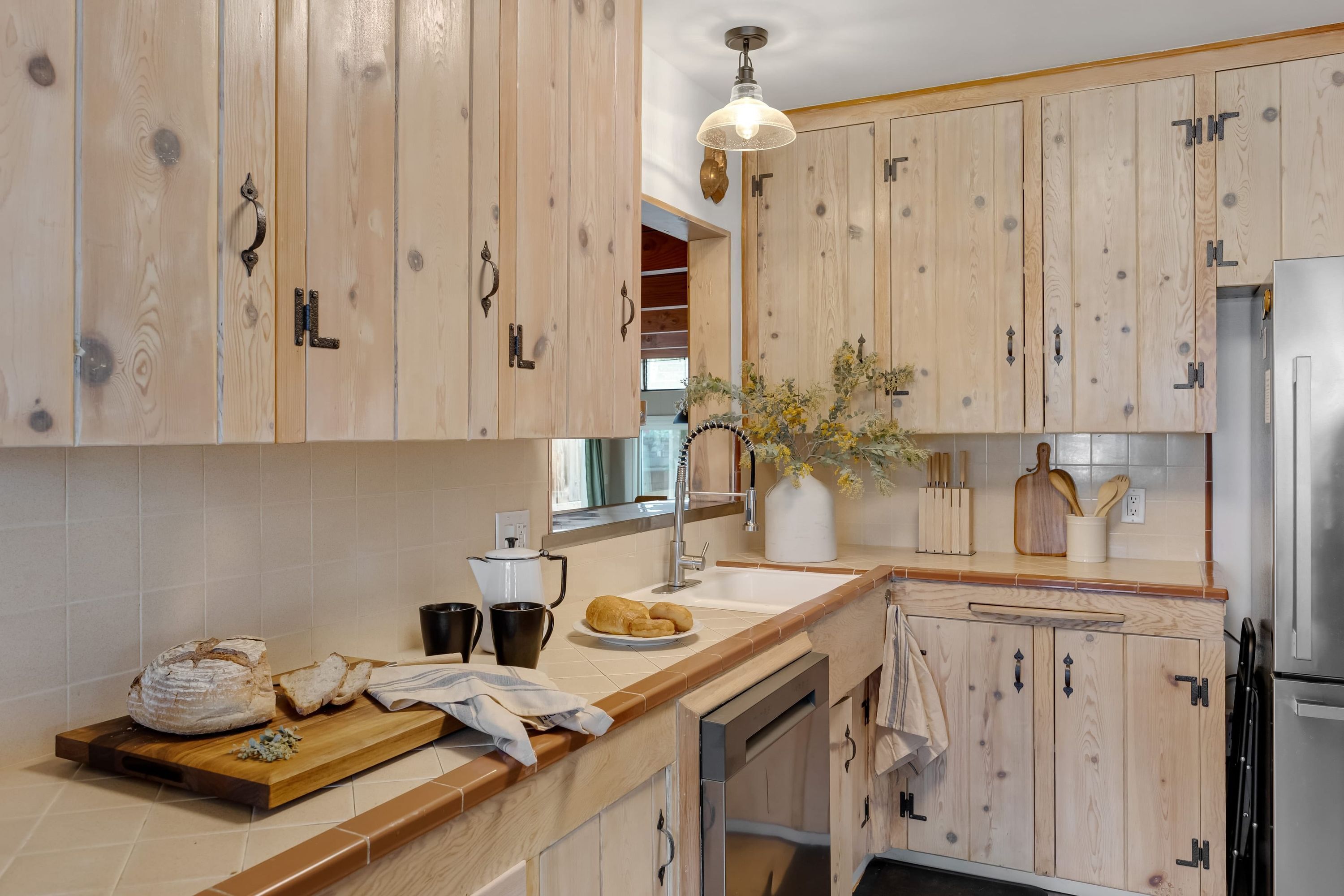 A kitchen with wooden cabinets, a stainless steel refrigerator, a wooden cutting board with bread, and a vase with flowers on the counter.