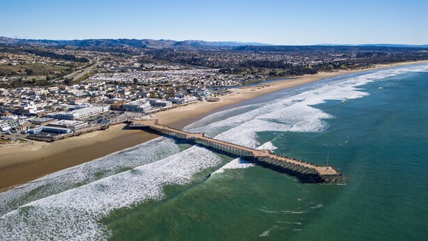 An aerial view of a coastal town with a long pier extending into the ocean, a sandy beach, and a densely built urban area in the background. - Casa do Sol by Avantstay Rooftop Deck w Ocean Views 2 Blocks to Beach (Pismo Beach)