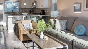 A modern living room with a sofa, a coffee table, and a plant. There is a kitchen in the background with white cabinets and a dining area with chairs. - Casa do Sol by Avantstay Rooftop Deck w Ocean Views 2 Blocks to Beach (Pismo Beach)