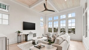 A modern living room with a white sofa, a coffee table with books and a plant, a TV mounted on the wall, and large windows offering a view of the outdoors. - Katydid Beach Bungalow by Avantstay Gulf Views New Construction in Seagrove (Santa Rosa Beach)