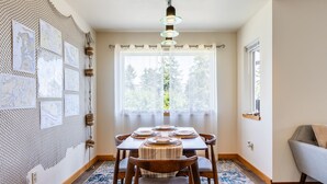 A dining area with a table set for four, a hanging light fixture, and a window with a view of trees. - Seaside Getaway by Avantstay Deck w Views (Langley)