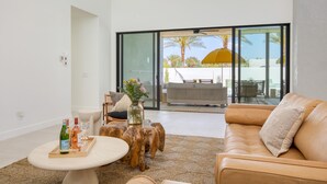 En moderne stue med en lædersofa, et rundt sofabord og et træsidebord. Der er en glasdør, der fører til en udendørs patio med en gul parasol.