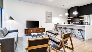 Ett modernt vardagsrum med en soffa, ett soffbord och stolar, en platt-TV, en köksdel med barstolar och ett stort fönster.