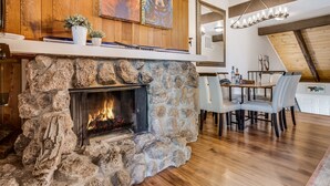 Un camino in pietra con fuoco acceso, pareti rivestite in legno, una zona pranzo con sedie e un lampadario.