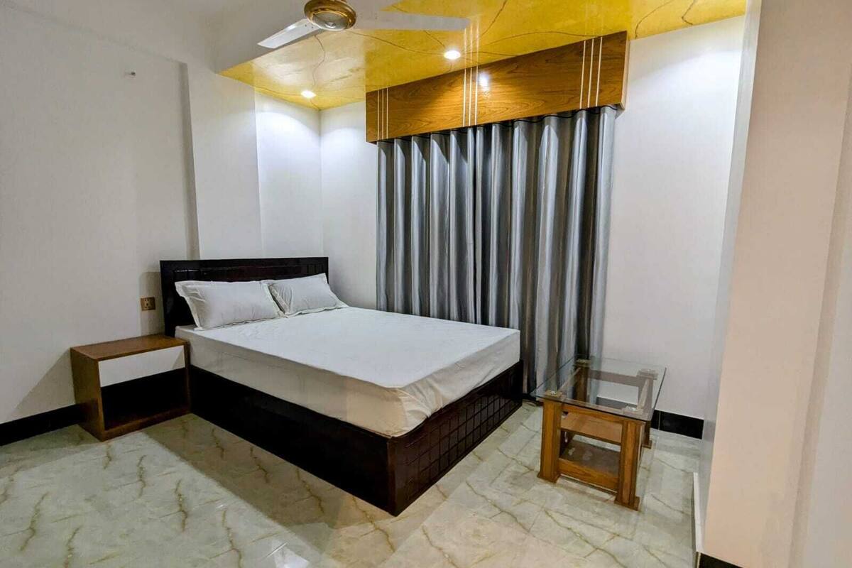 Une chambre à coucher comprenant un lit, une table de chevet, une petite table, un ventilateur de plafond et une fenêtre avec des rideaux.