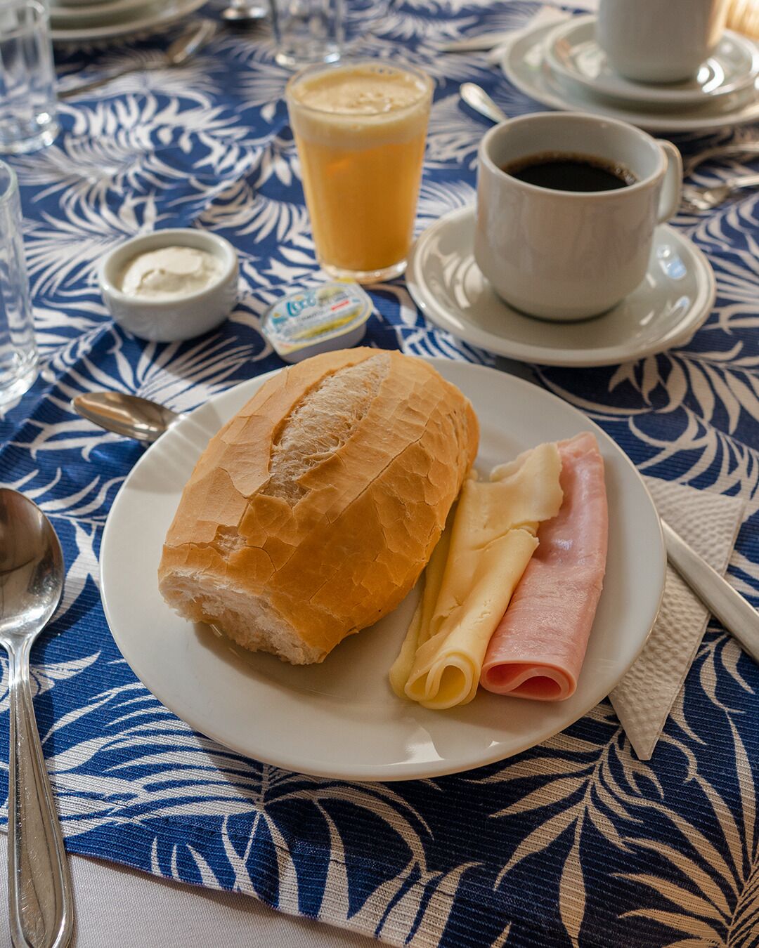A plate with a baguette and ham, a glass of orange juice, and a cup of coffee on a table with a blue and white pattern.
