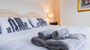 Een netjes opgemaakt bed met witte lakens en grijze handdoeken, een lamp op een nachtkastje en een lijst met een foto aan de muur.