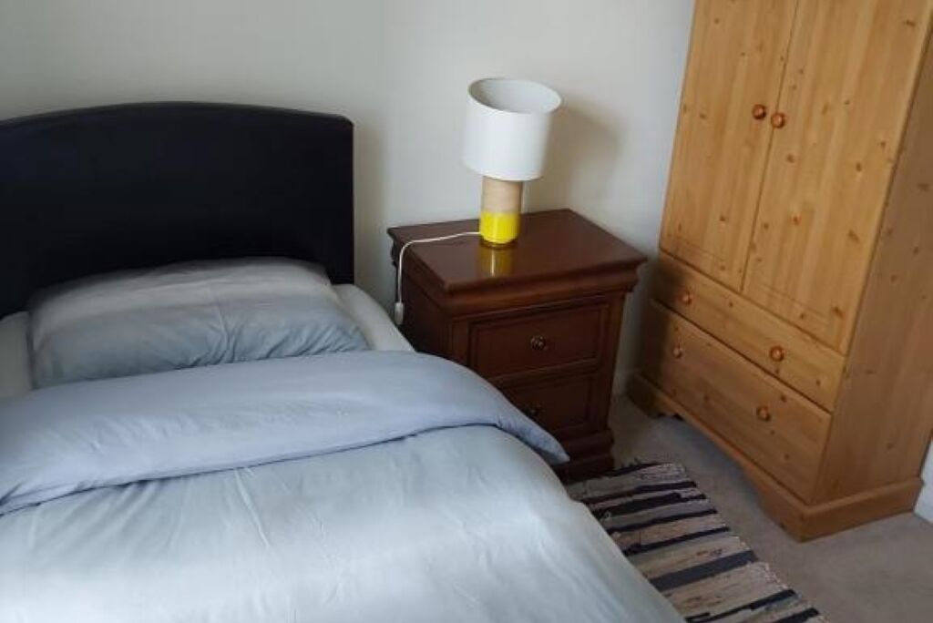 A bedroom with a bed, a nightstand with a lamp, a wooden wardrobe, and a striped rug.