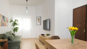 A living room with a green sofa, wooden coffee table, and a dining table with chairs. There is a TV on the wall, a plant, and a vase with flowers. - Elegantis Santa Croce Suite Apartment (Roma)