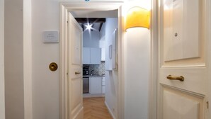 A hallway leading to a kitchen with white cabinets, a wooden floor, and a wall-mounted light fixture.
