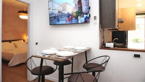 A room with a bed, a dining table set for two, a TV mounted on the wall, and a kitchen area with a window.