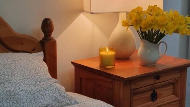Ein Bett mit einem gemusterten Überwurf, ein Nachttisch mit Kerze und eine Vase mit gelben Blumen.