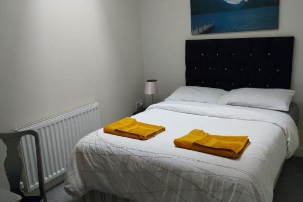 Een hotelkamer met een bed, wit beddengoed, een radiator, een lamp en een landschapsschilderij.