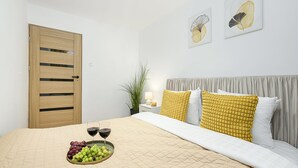 Un dormitorio con una puerta de madera, una cama con colcha color beige, una bandeja de uvas y vino, y dos cuadros en la pared.