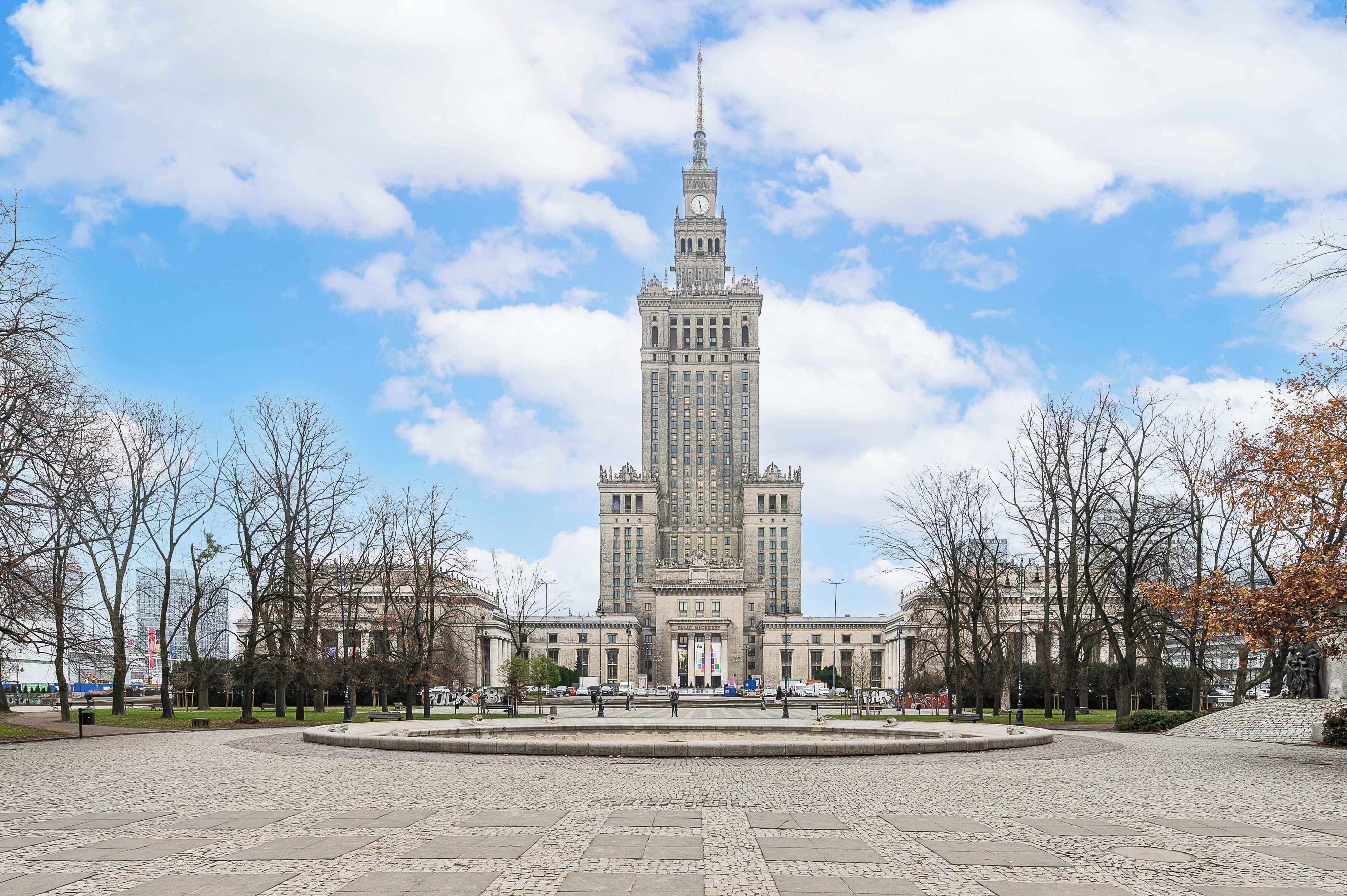 A large, historic building with a central clock tower, surrounded by a paved plaza and leafless trees.