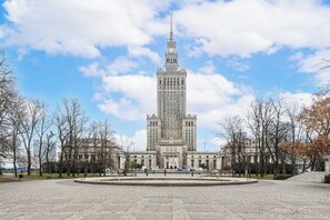 Un grande edificio storico con una torre dell'orologio centrale, circondato da una piazza lastricata e alberi spogli.