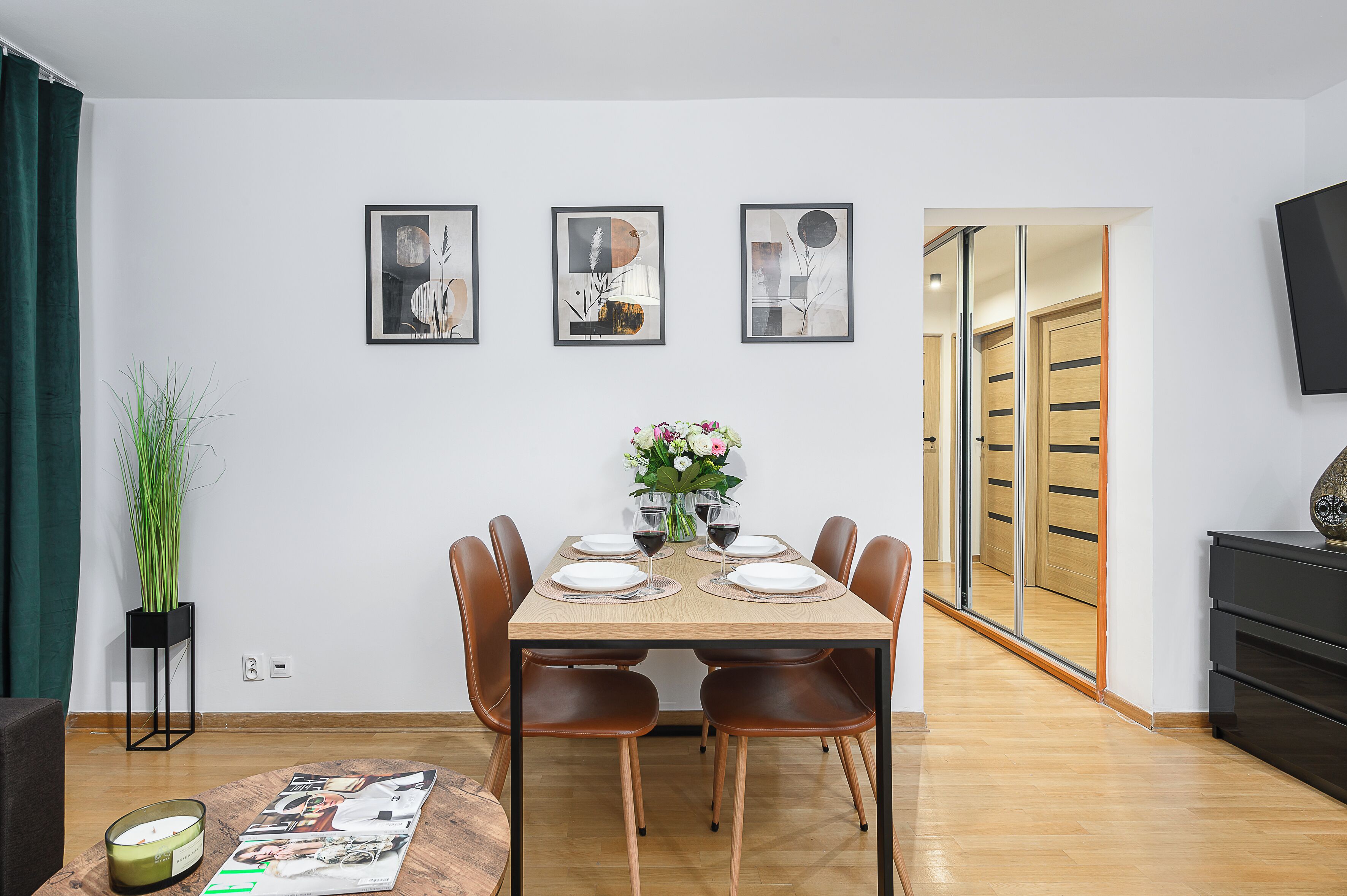 Ein Essbereich mit einem Holztisch, gedeckt für sechs Personen, umgeben von braunen Stühlen. An der Wand über dem Tisch hängen drei gerahmte Kunstwerke.