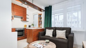 Una cocina moderna con armarios de madera y horno empotrado, un sofá gris oscuro con cojines, una mesa de centro redonda con una revista, y un ventanal con cortina verde.