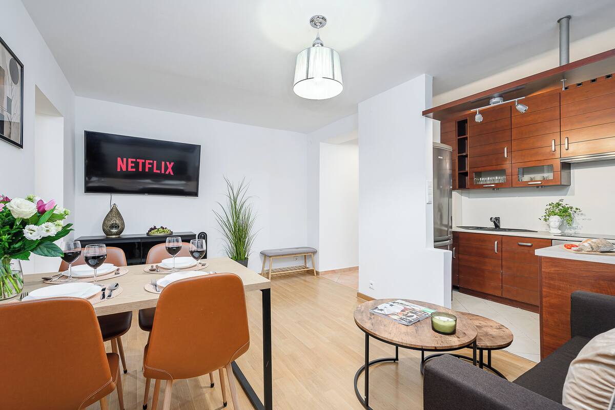 Un salon moderne comprenant un coin repas, une cuisine avec des meubles en bois, un téléviseur à écran plat et un banc.