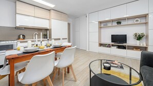 Una cocina moderna con armarios blancos, un comedor con mesa de madera y sillas blancas, y una sala de estar con sofá negro y una mesa de centro de vidrio.